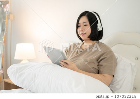 A beautiful Asian plus-size woman wearing headphones and using her tablet while relaxing on her bed. A beautiful Asian plus-size woman wearing headphones and using her tablet while relaxing on her bed. 107052218