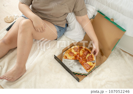 Cropped top view image of a curvy plus-size woman in comfy clothes eating pizza in her bedroom. Cropped top view image of a curvy plus-size woman in comfy clothes eating pizza in her bedroom. 107052246