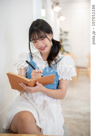 An attractive Asian woman in a cute dress is keeping her diary while relaxing in a coffee shop. An attractive Asian woman in a cute dress is keeping her diary while relaxing in a coffee shop. 107052286