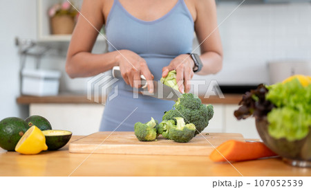 A fit woman in gym clothes is cutting broccoli on a chopping board, preparing her healthy breakfast. 107052539