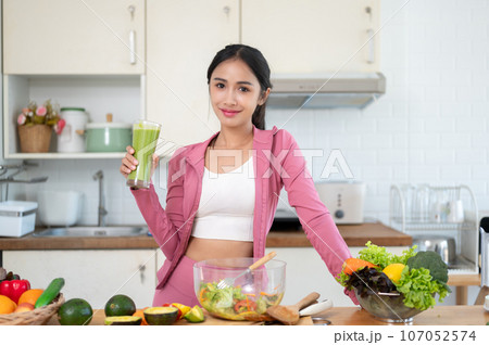 A slim Asian woman in gym clothes is holding a glass of a healthy green smoothie at a kitchen table. 107052574