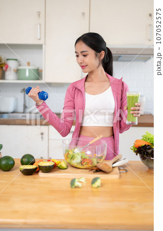 A slim Asian woman in sportswear is holding a dumbbell and a glass of green smoothie in the kitchen. 107052575