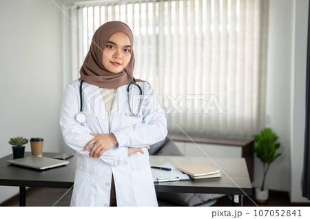 An attractive Asian Muslim female doctor in a uniform stands with her arms crossed in her office. An attractive Asian Muslim female doctor in a uniform stands with her arms crossed in her office. 107052841