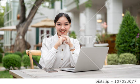 A beautiful Asian woman sits at a table with a laptop and her palms on her chin in her backyard. A beautiful Asian woman sits at a table with a laptop and her palms on her chin in her backyard. 107053077