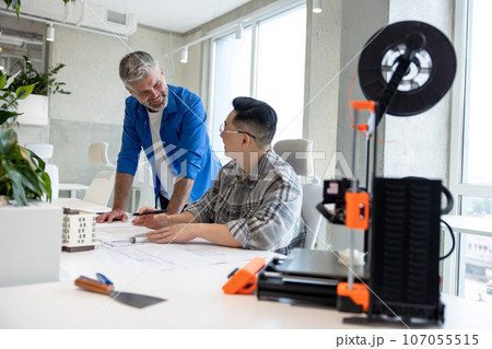 Men working with 3D printer and blueprint for designing mechanical parts in high tech workshop. 107055515