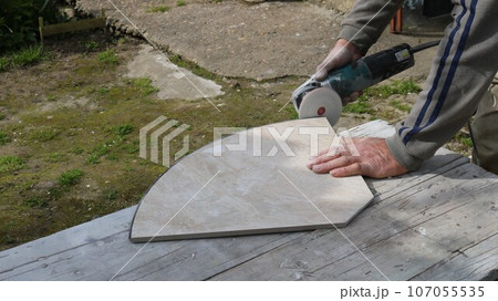 sanding the edge of a large decorative light tile in an outdoor workshop using a round sanding attachment, processing building finishing material with an angle saw with sandpaper sanding the edge of a large decorative light tile in an outdoor workshop using a round sanding attachment, processing building finishing material with an angle saw with sandpaper 107055535