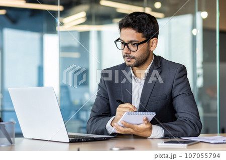 Serious and concentrated thinking businessman watching webinar online course, man upgrading at workplace inside office, hispanic man taking notes and writing data in notebook. Serious and concentrated thinking businessman watching webinar online course, man upgrading at workplace inside office, hispanic man taking notes and writing data in notebook. 107055794