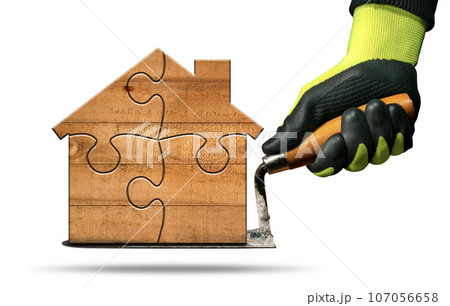 Gloved Hand Holding a Trowel and a Small Wooden Model House 107056658