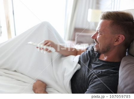 Smiling man lies in bed and presses buttons on remote control Smiling man lies in bed and presses buttons on remote control 107059785