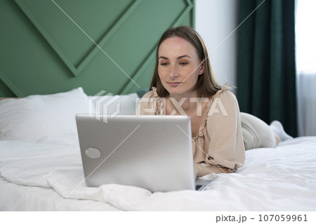 Beautiful young brunette woman working at home on a laptop lying on the bed Beautiful young brunette woman working at home on a laptop lying on the bed 107059961