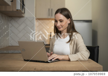 Young woman working online in her kitchen with a laptop Young woman working online in her kitchen with a laptop 107059969