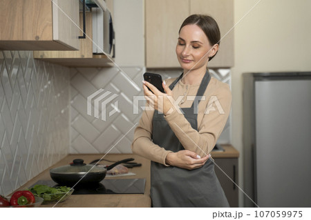 Young woman using a smartphone is studying at the kitchen table. A smiling woman reads a phone message. Brunette happy girl typing a text message Young woman using a smartphone is studying at the kitchen table. A smiling woman reads a phone message. Brunette happy girl typing a text message 107059975