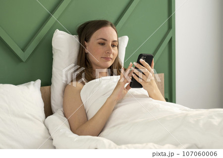 Portrait of a beautiful young brunette woman using a smart mobile phone on a bed with a white pillow-blanket in the bedroom interior Portrait of a beautiful young brunette woman using a smart mobile phone on a bed with a white pillow-blanket in the bedroom interior 107060006
