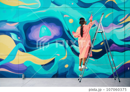 Female street artist painting colorful graffiti standing on a ladder using paint roller 107060375