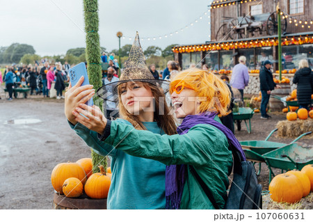 Two laughing female friends in fun glasses and witch hat having fun and making selfie on phone on decorated pumpkin farm. Selecting Thanksgiving and Halloween holidays decor. Autumn fall festive mood 107060631