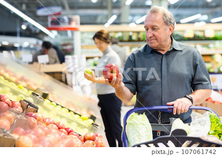 Elderly man chooses apples in vegetable and fruit department Elderly man chooses apples in vegetable and fruit department 107061742