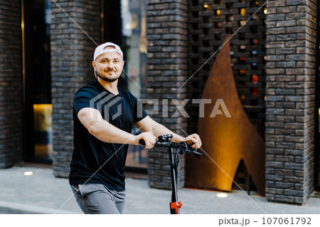 Young handsome man in white baseball cap riding on scooter to work along the street in the city Young handsome man in white baseball cap riding on scooter to work along the street in the city 107061792