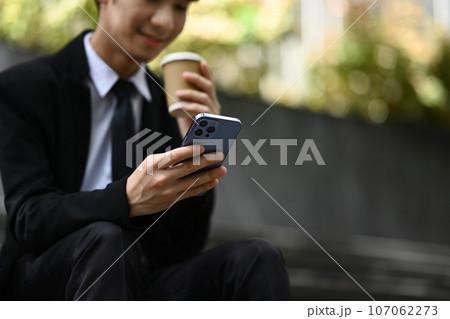 Pleasant businessman texting messages, chatting in social on smartphone while sitting on stairs outside office building Pleasant businessman texting messages, chatting in social on smartphone while sitting on stairs outside office building 107062273