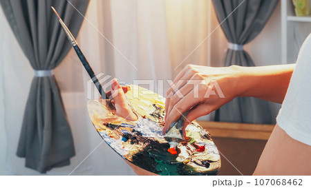 Art process. Creating artwork. Closeup of woman painter hand mixing oil colorful paint on messy palette with paintbrush at artist studio. 107068462