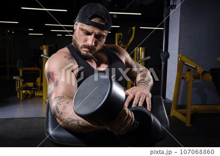 Tattooed, bearded guy in black vest and cap. He is lifting a dumbbell, training his biceps, sitting on preacher curl bench at dark gym. Close up Tattooed, bearded guy in black vest and cap. He is lifting a dumbbell, training his biceps, sitting on preacher curl bench at dark gym. Close up 107068860