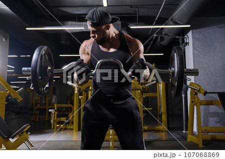 Tattooed male in black sport gloves, shorts, vest and cap. Lifting barbell, training his biceps, standing in dark gym with yellow equipment. Close up 107068869