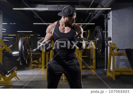Sportsman in black sport gloves, shorts, vest and cap. He is lifting barbell, training his biceps, posing in dark gym with yellow equipment. Close up 107068885