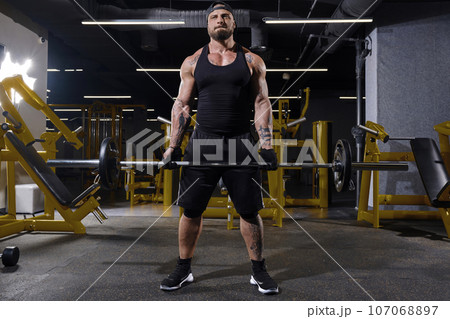 Bearded athlete in black sport gloves, shorts, vest, cap and sneakers. He lifting a barbell, posing in dark gym with yellow equipment. Full length 107068897