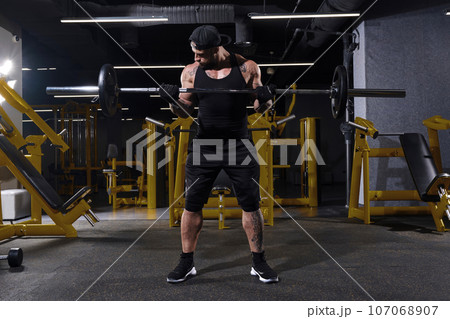 Bearded sportsman in black sport gloves, shorts, vest, cap and sneakers. He lifting a barbell, posing in dark gym with yellow equipment. Full length Bearded sportsman in black sport gloves, shorts, vest, cap and sneakers. He lifting a barbell, posing in dark gym with yellow equipment. Full length 107068907