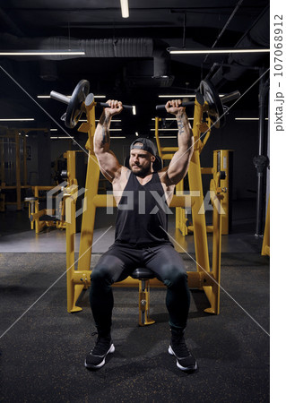 Tattooed, bearded male in black sweatpants, vest and cap. He performing a chest press while sitting on an exercise machine at dark gym. Full length 107068912