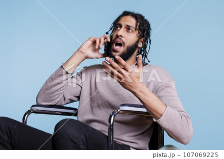 Emotional irritated arab man with disability talking on smartphone while sitting in wheelchair. Angry person with physical imparment making gestures while chatting on mobile phone 107069225