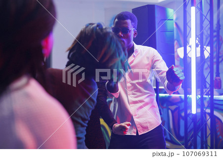 Cool african american man partying and dancing in nightclub illuminated with vibrant lights. Clubbers having fun on dancefloor in crowded club while attending discotheque 107069311
