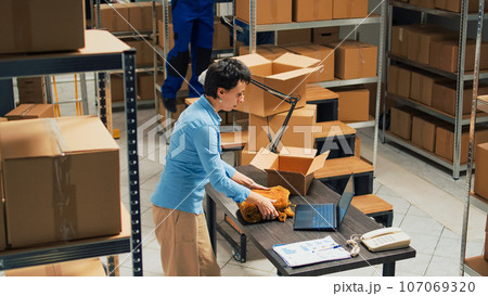 Woman employee taking supplies from warehouse shelves and putting them in cardboard boxes, using packages for delivery. Young adult writing inventory information on laptop, logistics. 107069320