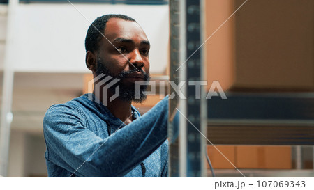 African american worker using scanner and tablet to check stock inventory, working on products management. Young man scanning merchandise in cardboard boxes on warehouse shelves. Handheld shot. African american worker using scanner and tablet to check stock inventory, working on products management. Young man scanning merchandise in cardboard boxes on warehouse shelves. Handheld shot. 107069343