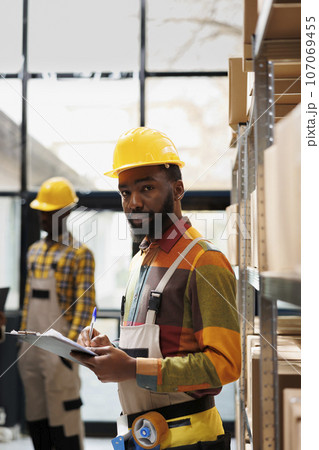 African american logistics supervisor doing merchandise inventory in warehouse portrait. Distribution manager taking notes on clipboard about products supply chain and looking at camera African american logistics supervisor doing merchandise inventory in warehouse portrait. Distribution manager taking notes on clipboard about products supply chain and looking at camera 107069455