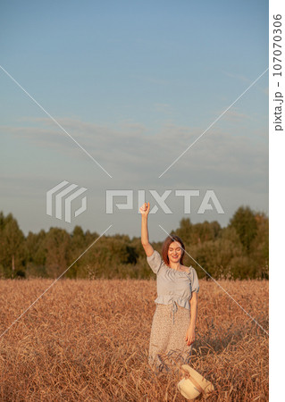 A young beautiful girl with braces on her teeth and long hair poses in a wheat field in the summer at sunset. The girl holds a hat in her hand against the background of a wheat field. A young beautiful girl with braces on her teeth and long hair poses in a wheat field in the summer at sunset. The girl holds a hat in her hand against the background of a wheat field. 107070306