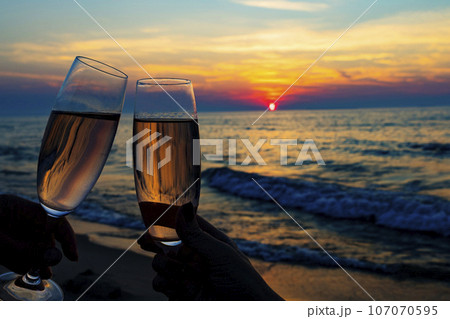 close up of two white wine glasses cheering with beach sunset background 107070595