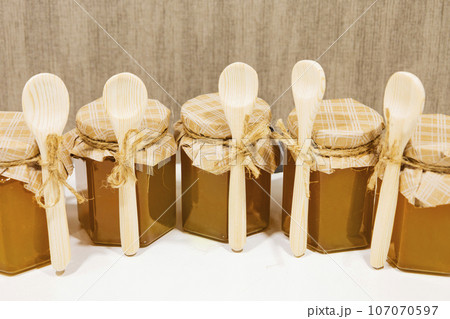 A small yellow jar of honey with a decorative lid made of beige paper and a bow, a small wooden spoon A small yellow jar of honey with a decorative lid made of beige paper and a bow, a small wooden spoon 107070597