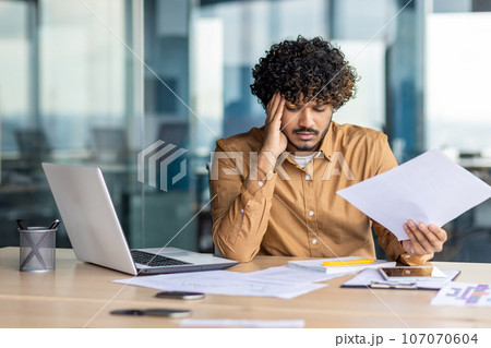 Upset financier looking at sales and revenue chart of company, businessman inside office disappointed with sales results and achievements, Hispanic man sitting at table with documents and laptop. 107070604