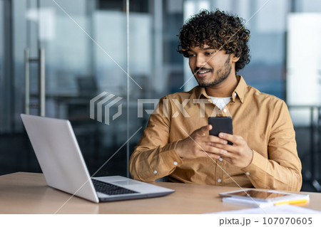 Young successful hispanic businessman using app on phone, man smiling contentedly inside office at workplace, employee working with laptop. 107070605