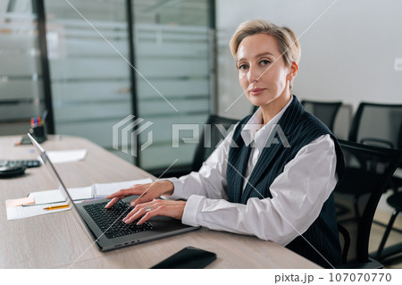 Portrait of elegance middle-aged businesswoman executive top manager sitting at desk working with documents typing on laptop computer, looking at camera with serious expression, in corporate office. Portrait of elegance middle-aged businesswoman executive top manager sitting at desk working with documents typing on laptop computer, looking at camera with serious expression, in corporate office. 107070770