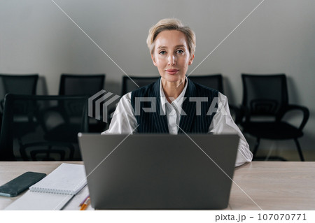 Front view of elegance middle-aged businesswoman executive top manager sitting at desk working with documents typing on laptop computer, looking at camera with serious expression, in corporate office. 107070771