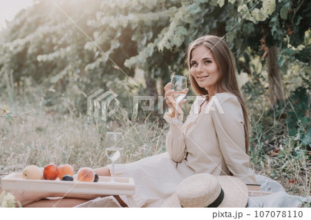 Woman picnic vineyard. Happy woman with a glass of wine at a picnic in the vineyard, wine tasting at sunset and open nature in the summer. Romantic dinner, fruit and wine. 107078110