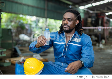 Man African American engineer holding plastic bottle drinking water  after working at factory. 107080102