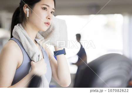 スポーツジムで汗を拭くワイヤレスイヤホンをした若い女性 107082622