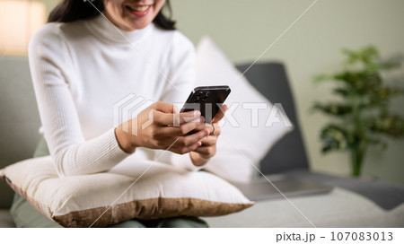 Cropped image of a beautiful young Asian woman using her smartphone while sitting on a couch 107083013