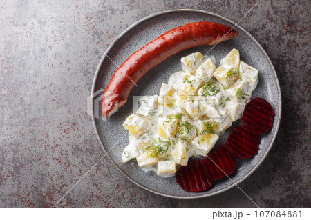 Lightly smoked sausage with a side dish of potatoes with dill and boiled beets close-up on a plate on the table. Horizontal top view 107084881