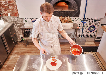 Cooking pizza. the workpiece poured tomato sauce. Closeup hand of chef baker in uniform apron cook at kitchen Cooking pizza. the workpiece poured tomato sauce. Closeup hand of chef baker in uniform apron cook at kitchen 107086881