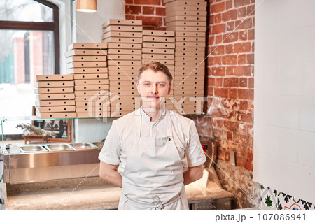 The chef prepares pizza in a neapolitan pizzeria. Cook in a apron in the kitchen with a shovel in his hands. boxes for food delivery on background. 107086941