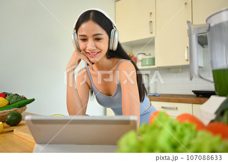 Happy young woman making detox vegetable smoothie and following online recipe on digital tablet. Dieting concept Happy young woman making detox vegetable smoothie and following online recipe on digital tablet. Dieting concept 107088633