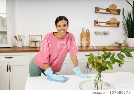 Happy smiling girl is cleaning the house, washing the surface of the dining table. Cute young woman in homemade clothes and rubber gloves wipes dust from the kitchen table. Cleaning with rag. 107089279